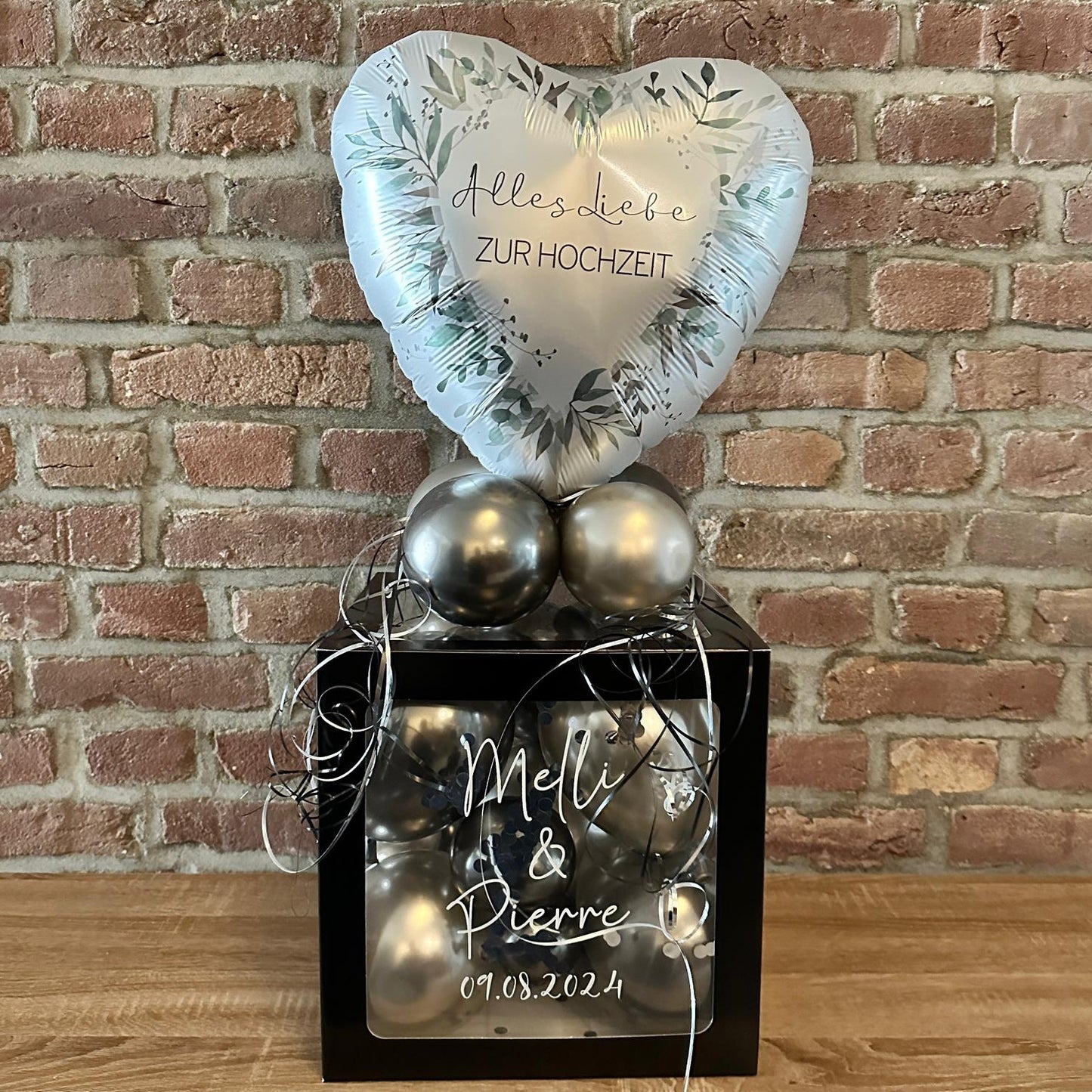 Geschenkbox schwarz mit weißer Aufschrift und silbernen Ballons gefüllt.